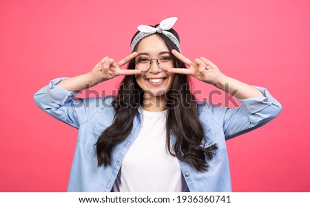 Funny cheerful asian teenage girl, with long black hair, in a denim shirt and round glasses, shows a sign of peace near the eyes, calls for peace, against war, posing on a pink background.