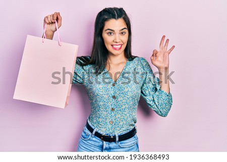 Young hispanic girl holding shopping bags doing ok sign with fingers, smiling friendly gesturing excellent symbol 