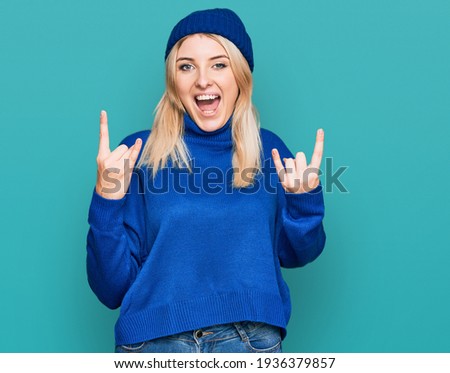 Young caucasian woman wearing wool winter sweater and cap shouting with crazy expression doing rock symbol with hands up. music star. heavy concept. 