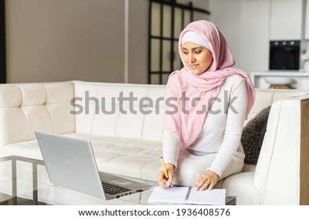 Portrait of a young muslim woman in hijab working at home using laptop computer while sitting on sofa, writing notes, writes a to-do list, working remotely, studying online, freelancer writing details