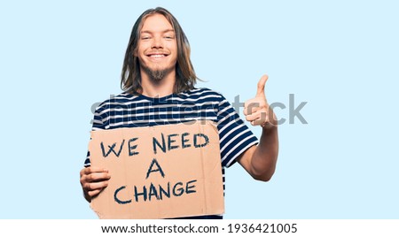 Handsome caucasian man with long hair holding we need a change banner smiling happy and positive, thumb up doing excellent and approval sign 