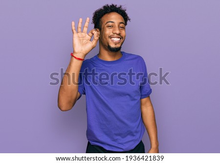 Young african american man with beard wearing casual purple t shirt smiling positive doing ok sign with hand and fingers. successful expression. 