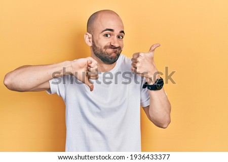Young bald man wearing casual white t shirt doing thumbs up and down, disagreement and agreement expression. crazy conflict 