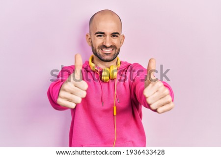 Young bald man wearing gym clothes and using headphones approving doing positive gesture with hand, thumbs up smiling and happy for success. winner gesture. 
