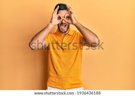Young hispanic man wearing casual yellow t shirt doing ok gesture like binoculars sticking tongue out, eyes looking through fingers. crazy expression. 