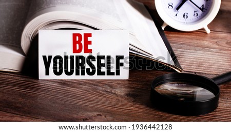 The words BE YOURSELF written on a white card near an open book, alarm clock and magnifying glass