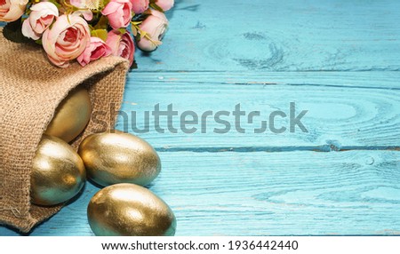    Golden Easter eggs in jute bag and beautiful flowers on blue rustic old wooden background. Trendy Easter composition. Copy space for text. Minimal Easter concept. Banner.                           