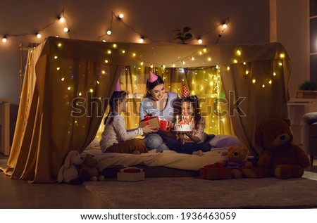 Family time. Two twin sisters with their mother are sitting in a cozy tent bed and celebrating their birthday. Family in holiday hats exchanges gifts and gathers to blow out the candles on the cake.