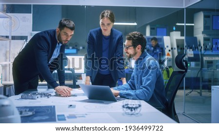 Modern Factory Office Meeting Room: Diverse Team of Engineers, Managers Talking at Conference Table, Look at Blueprints, Inspect Mechanism, Use Laptop. High-Tech Facility with CNC Machines Royalty-Free Stock Photo #1936499722