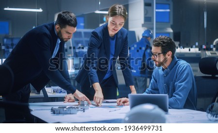 Modern Factory Office Meeting Room: Diverse Team of Engineers, Managers Talking at Conference Table, Look at Blueprints, Inspect Mechanism, Use Laptop. High-Tech Facility with CNC Machines Royalty-Free Stock Photo #1936499731