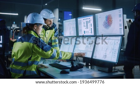 Industry 4.0 Modern Factory: Project Engineer Talks to Female Operator who Controls Facility Production Line, Uses Computer with Screens Showing AI, Machine Learning Enhanced Assembly Process Royalty-Free Stock Photo #1936499776