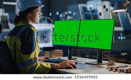 Young and Confident Industrial Female Engineer in Safety Vest and Hardhat is Working on Computer with Green Screen Mock-up Template. Industrial Factory with High-Tech CNC Machinery