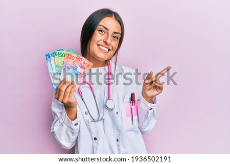 Beautiful hispanic woman wearing medical uniform holding swiss franc banknotes smiling happy pointing with hand and finger to the side 