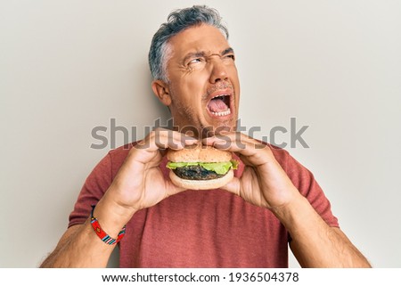 Handsome middle age mature man eating a tasty classic burger angry and mad screaming frustrated and furious, shouting with anger. rage and aggressive concept.  Royalty-Free Stock Photo #1936504378