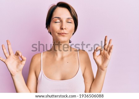 Young brunette woman with short hair over pink background relax and smiling with eyes closed doing meditation gesture with fingers. yoga concept. 