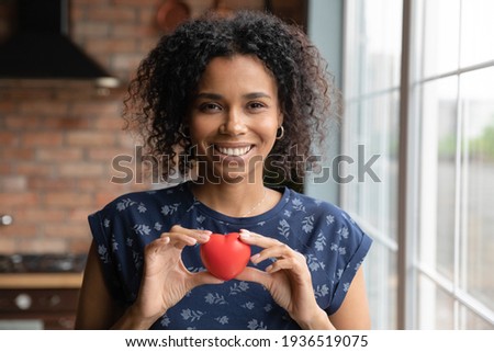 Headshot portrait of happy millennial black woman looking at camera holding small red toy heart in hands at breast height. Smiling young mixed race female express gratitude appreciation on photo video