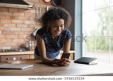 In a good mood. Happy young biracial woman stand by table near large kitchen window texting messaging online using cell device. Smiling black teen female spend time surfing web on modern smartphone Royalty-Free Stock Photo #1936519234