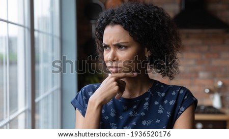 Pensive millennial african female having bad day stand by window lost in unpleasant thoughts. Angry unhappy young black lady frowning look at distance rub chin ponder on way out of hard life situation Royalty-Free Stock Photo #1936520176