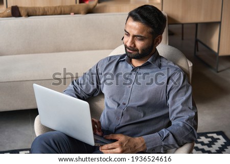 Serious bearded indian business man working on laptop from home office sitting in chair. Male indian entrepreneur using computer remote studying, browsing web, having virtual meeting, watching webinar
