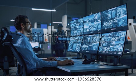 Industry 4.0 Modern Factory: Security Operator Controls Proper Functioning of Workshop Production Line, Uses Computer with Screens Showing Surveillance Camera Feed. High-Tech Security Royalty-Free Stock Photo #1936528570