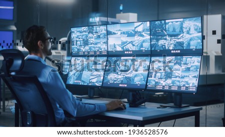 Industry 4.0 Modern Factory: Security Operator Controls Proper Functioning of Workshop Production Line, Uses Computer with Screens Showing Surveillance Camera Feed. High-Tech Security Royalty-Free Stock Photo #1936528576