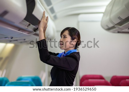 Young beautiful Asian airline cabin crew attendant closing overhead cabinet luggage compartment for checking safety of passenger before flight. Airline business professional uniform occupation concept Royalty-Free Stock Photo #1936529650