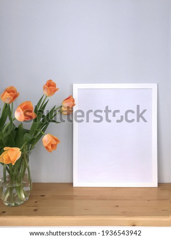 A white empty frame on the table for a photo or painting in the interior and delicate orange-pink tulips in a glass vase next to it