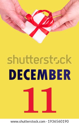 December 11th. Festive Vertical Calendar With Hands Holding White Gift Box With Red Ribbon And Calendar Date 11 December On Illuminating Yellow Background. Winter month, day of the year concept.