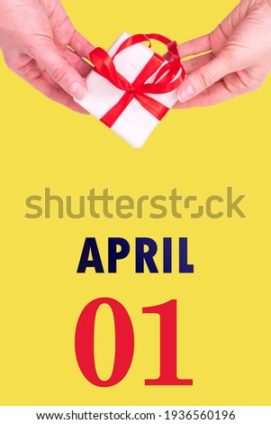 April 1st. Festive Vertical Calendar With Hands Holding White Gift Box With Red Ribbon And Calendar Date 1 April On Illuminating Yellow Background. Spring month, day of the year concept. Royalty-Free Stock Photo #1936560196