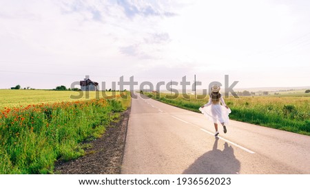 back view of girl in dress and boater walks on the road near the field of winter wheat and poppies. blue sky with clouds.