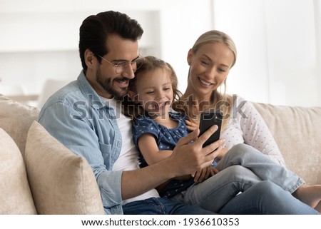 Happy young Caucasian family with small daughter relax on couch using smartphone together. Smiling mom and dad rest on sofa with little girl child talk on video call on cellphone gadget online.