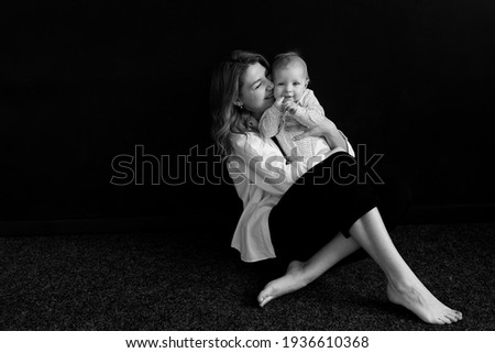 Happy successful woman sitting and holding adorable little girl in her arms. Caring mother gently hugging and kissing her sweet cute child, posing in studio, photoshoot concept