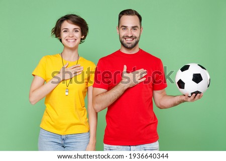 Smiling young couple friends sport family woman man football fans in yellow red t-shirts cheer up support favorite team with soccer ball put hands on chest hearts isolated on green background studio Royalty-Free Stock Photo #1936640344
