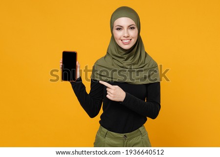 Smiling young arabian muslim woman in hijab black green clothes pointing index finger on mobile phone with blank empty screen isolated on yellow background studio. People religious lifestyle concept