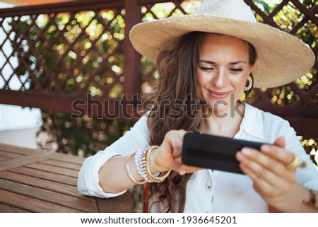smiling stylish female in white shirt with hat sending text message using smartphone in the terrace of guest house hotel.