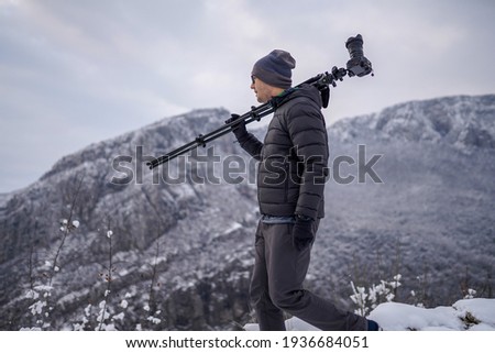Young male photographer carrying tripod with camera on shoulder while walking across mountain
