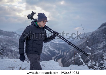 Photographer with eyeglasses carrying tripod with camera on shoulder while walking across mountain