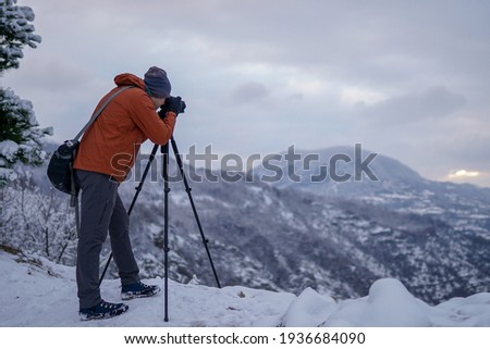 Male photographer standing on mountain viewpoint on snowy day and taking picture with camera on tripod