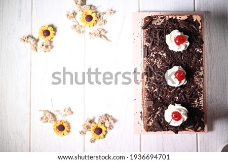 Black forest cake, decorated with whipped cream and cherries, on white wooden table with little yellow flower as tabel decoration. Selective focus. Top view.