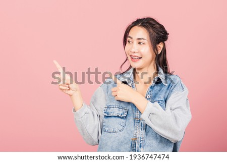 Asian happy portrait beautiful cute young woman wear denim standing pointing finger side away presenting product looking to side, studio shot isolated on pink background with copy space