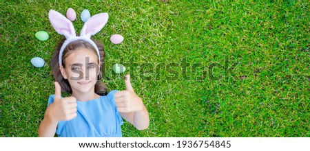 Happy girl wearing Easter bunny ears holds easter egg and lies on green summer grass ahd shows thumbs up gesture. Top down view. Empty space for text