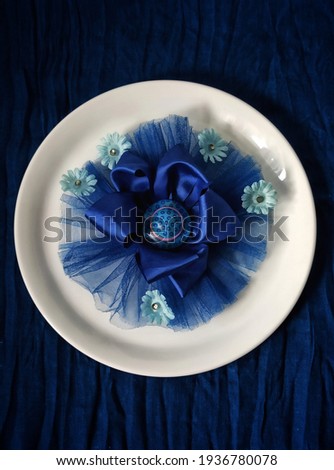 Selective focus on blue hair ribbon on white and glossy plate, in the blue blurry tangled cloth base. Kids Hair ribbon with blue small flower ornaments. Hair style. Girly.
