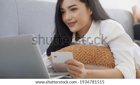 Asian woman holding credit card and using laptop to shopping online, lying on sofa. Shopaholic girl selecting products to cart in online website And pay via internet banking.