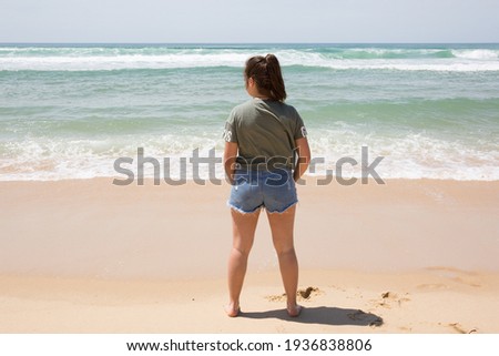 Girl in short jeans standing at ocean sand beach edge facing sea coast