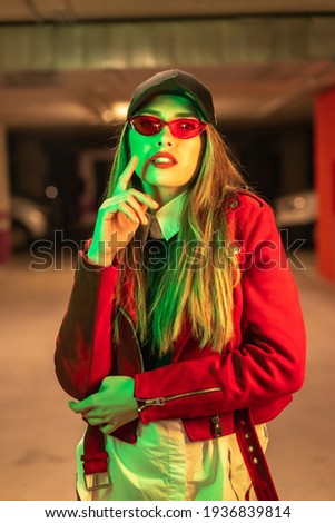 Photography with red and green neons in a parking lot. Portrait of a young pretty blond Caucasian woman in a red suit, sunglasses and a black cap
