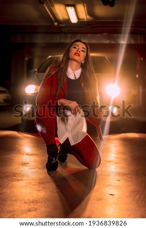 Photography with red neon in front of a car with the lights in a parking lot. Portrait of a pretty young blond Caucasian woman crouching