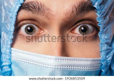 Shocked suprised amazed nurse doctor standing near wall holding his head. Male dressed in protective medical suit and blue face surgical mask. Coronavirus concept. Fear, fright, anxiety, alarm