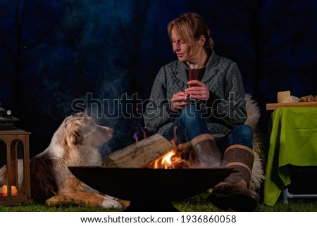 Young woman sitting by the campfire drinking wine. She watches her Australian Shepherd enjoy the winter weather. Outside in the park during blue hour