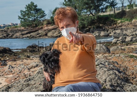 Portrait of senior woman with disposable medical mask and her dog on the beach pointing in a direction