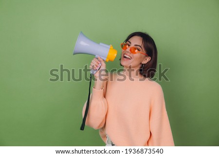 Young woman in casual peach and orange eyeglasses sweater isolated on green olive background happy screaming in megaphone copy space